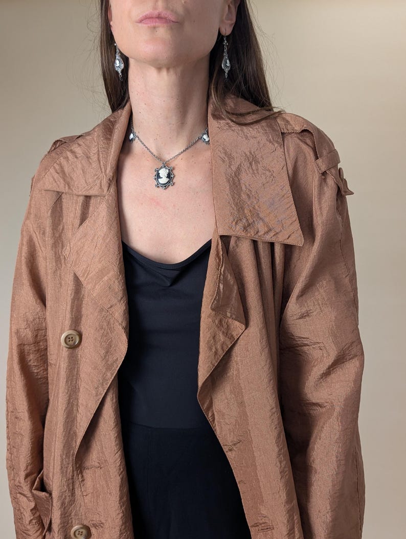 May include: A light brown trench coat with large lapels and two visible buttons. A black top is worn underneath. A silver necklace and earrings with cameo pendants are also visible.