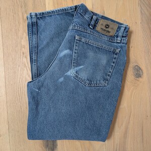 Op de afbeelding: Blauwe Wrangler jeans van denim, opgevouwen op een licht houten oppervlak. De jeans hebben een klassiek ontwerp met vijf zakken, een bruine leren patch met het Wrangler-logo en een vervaagde uitstraling. De jeans zijn van een medium wassing.