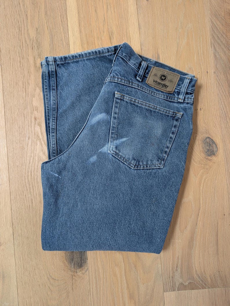 Op de afbeelding: Blauwe Wrangler jeans van denim, opgevouwen op een licht houten oppervlak. De jeans hebben een klassiek ontwerp met vijf zakken, een bruine leren patch met het Wrangler-logo en een vervaagde uitstraling. De jeans zijn van een medium wassing.