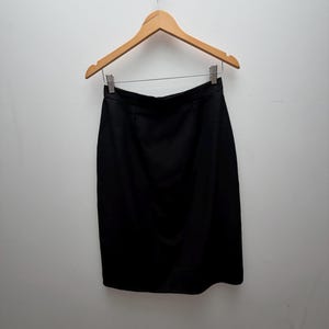 Puede incluir: Una falda negra lisa hasta la rodilla con una cintura estructurada y una silueta recta. La falda est&aacute; colgada de una percha de madera clara que cuenta con dos clips de metal plateado que sujetan firmemente el borde superior de la prenda.