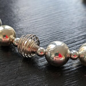 Peut inclure: Gros plan sur un collier argent&eacute; compos&eacute; de grosses perles sph&eacute;riques et d'un &eacute;l&eacute;ment en spirale unique. Les perles refl&egrave;tent la lumi&egrave;re, cr&eacute;ant un aspect brillant. Le collier est pr&eacute;sent&eacute; sur une surface sombre.