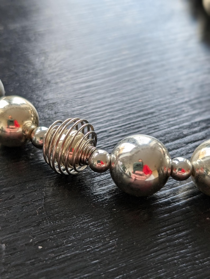 Peut inclure: Gros plan sur un collier argent&eacute; compos&eacute; de grosses perles sph&eacute;riques et d'un &eacute;l&eacute;ment en spirale unique. Les perles refl&egrave;tent la lumi&egrave;re, cr&eacute;ant un aspect brillant. Le collier est pr&eacute;sent&eacute; sur une surface sombre.