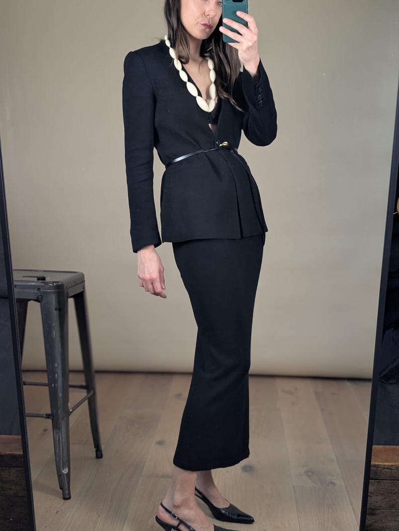 May include: A black skirt suit with a matching blazer and a pencil skirt. The blazer is cinched at the waist with a thin black belt. A white beaded necklace adds contrast. The outfit is paired with black heels.