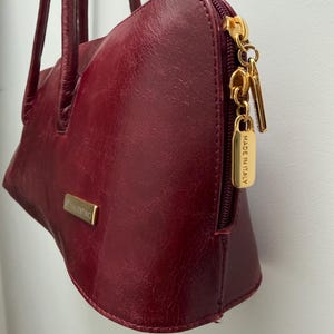 May include: A burgundy leather handbag with a gold zipper and hardware. The bag has a rectangular gold plate with the name "VALENTINO" on it. The zipper pull has a small gold tag that says "MADE IN ITALY".