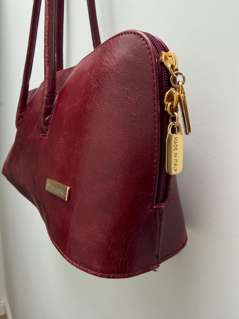 May include: A burgundy leather handbag with a gold zipper and hardware. The bag has a rectangular gold plate with the name "VALENTINO" on it. The zipper pull has a small gold tag that says "MADE IN ITALY".