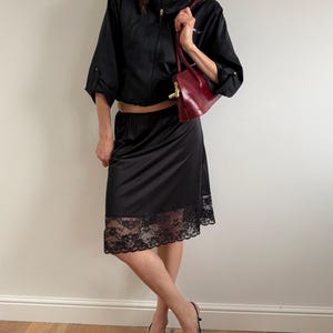 May include: A black outfit consisting of a cropped zip-up jacket, a slip skirt with a lace hem, and pointed-toe heels. A burgundy handbag with a gold clasp is held in the hand.
