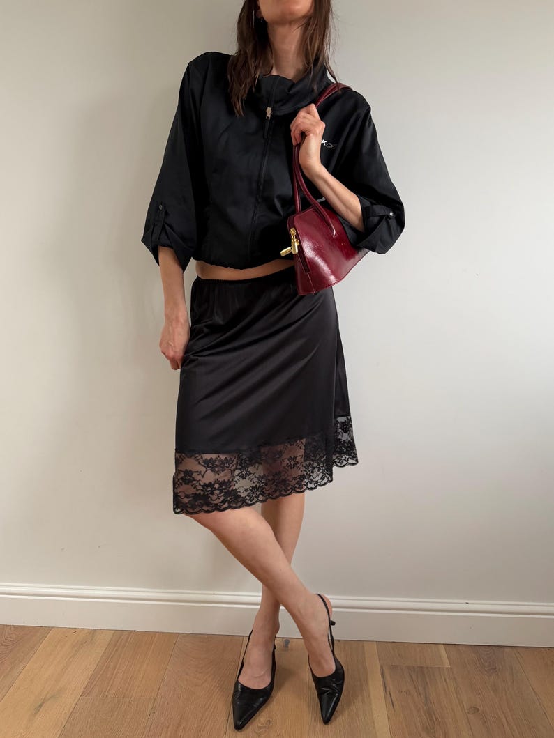 May include: A black outfit consisting of a cropped zip-up jacket, a slip skirt with a lace hem, and pointed-toe heels. A burgundy handbag with a gold clasp is held in the hand.