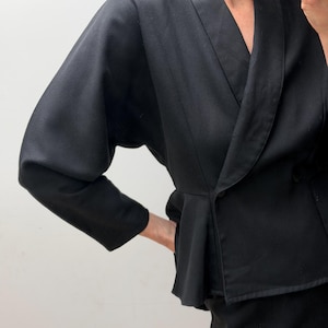 Puede incluir: Un blazer negro liso con un cuello chal ancho, mangas largas de forma ligeramente voluminosa y una cintura estilo peplum. La chaqueta tiene un cierre de un solo bot&oacute;n en la parte delantera y un corte entallado que se ensancha ligeramente en las caderas.