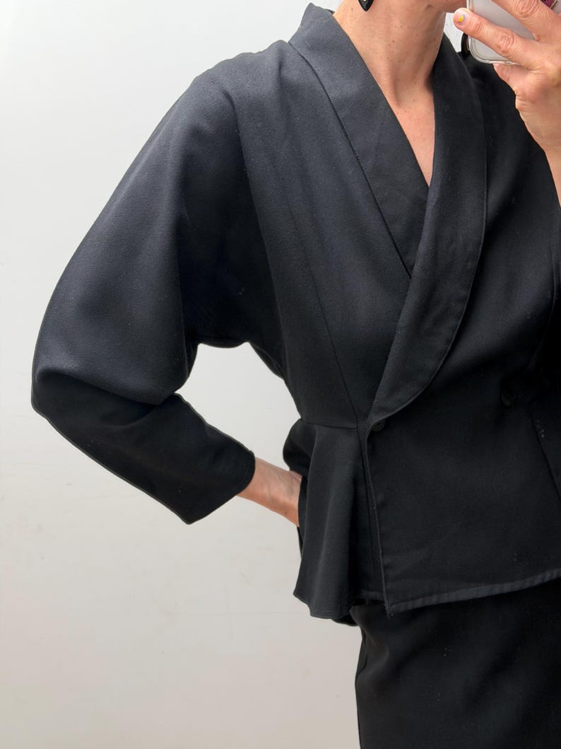 Puede incluir: Un blazer negro liso con un cuello chal ancho, mangas largas de forma ligeramente voluminosa y una cintura estilo peplum. La chaqueta tiene un cierre de un solo bot&oacute;n en la parte delantera y un corte entallado que se ensancha ligeramente en las caderas.