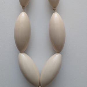 May include: Cream-colored necklace featuring large, oval beads strung together. The beads have a glossy finish and are connected by a thin, neutral-colored cord. The necklace is displayed against a plain, light background.