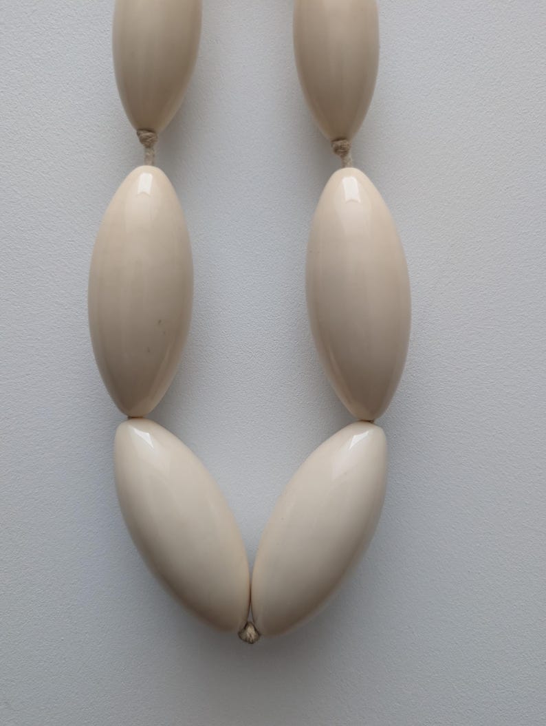 May include: Cream-colored necklace featuring large, oval beads strung together. The beads have a glossy finish and are connected by a thin, neutral-colored cord. The necklace is displayed against a plain, light background.
