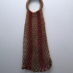 Könnte beinhalten: Eine gewebte Netztasche mit rundem Griff. Die Tasche weist ein Muster aus abwechselnd braunen und roten vertikalen Streifen auf. Die Tasche besteht aus einem Naturfasermaterial.