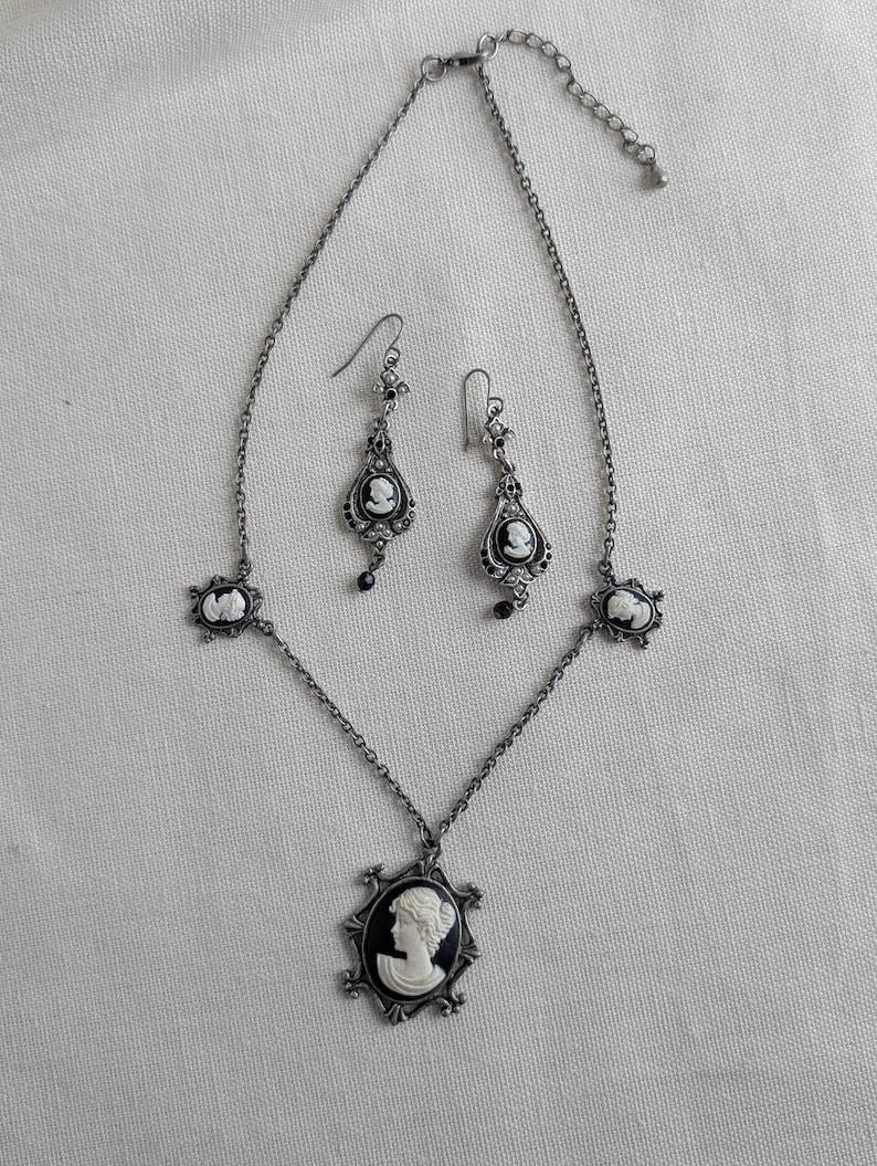 May include: A vintage-style jewelry set featuring a necklace and matching earrings. The set is silver-toned with black and white cameo pendants. The necklace has a large central pendant and smaller side pendants. The earrings have a teardrop shape.