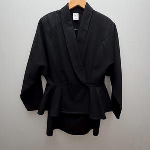 Puede incluir: Una chaqueta negra de manga larga con cuello tipo chal, cierre frontal cruzado con un solo bot&oacute;n visible y cintura con volante tipo peplum. La prenda se muestra colgada en una percha de madera sobre un fondo liso de color claro.