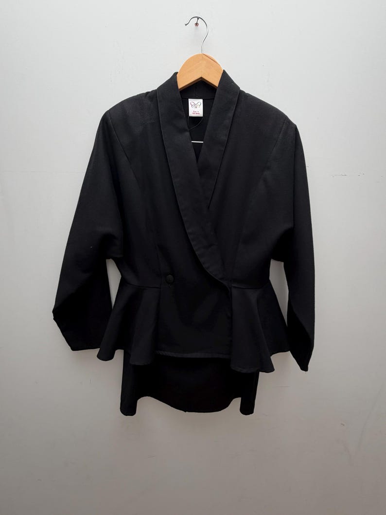 Puede incluir: Una chaqueta negra de manga larga con cuello tipo chal, cierre frontal cruzado con un solo bot&oacute;n visible y cintura con volante tipo peplum. La prenda se muestra colgada en una percha de madera sobre un fondo liso de color claro.