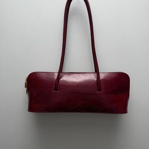 May include: A burgundy leather handbag with a rectangular shape and two long, arched handles. The bag has a gold-colored zipper and a glossy finish, suitable for carrying essentials.