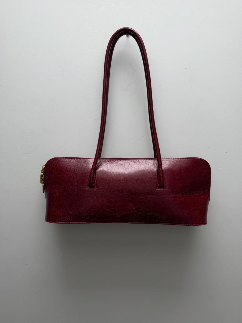 May include: A burgundy leather handbag with a rectangular shape and two long, arched handles. The bag has a gold-colored zipper and a glossy finish, suitable for carrying essentials.