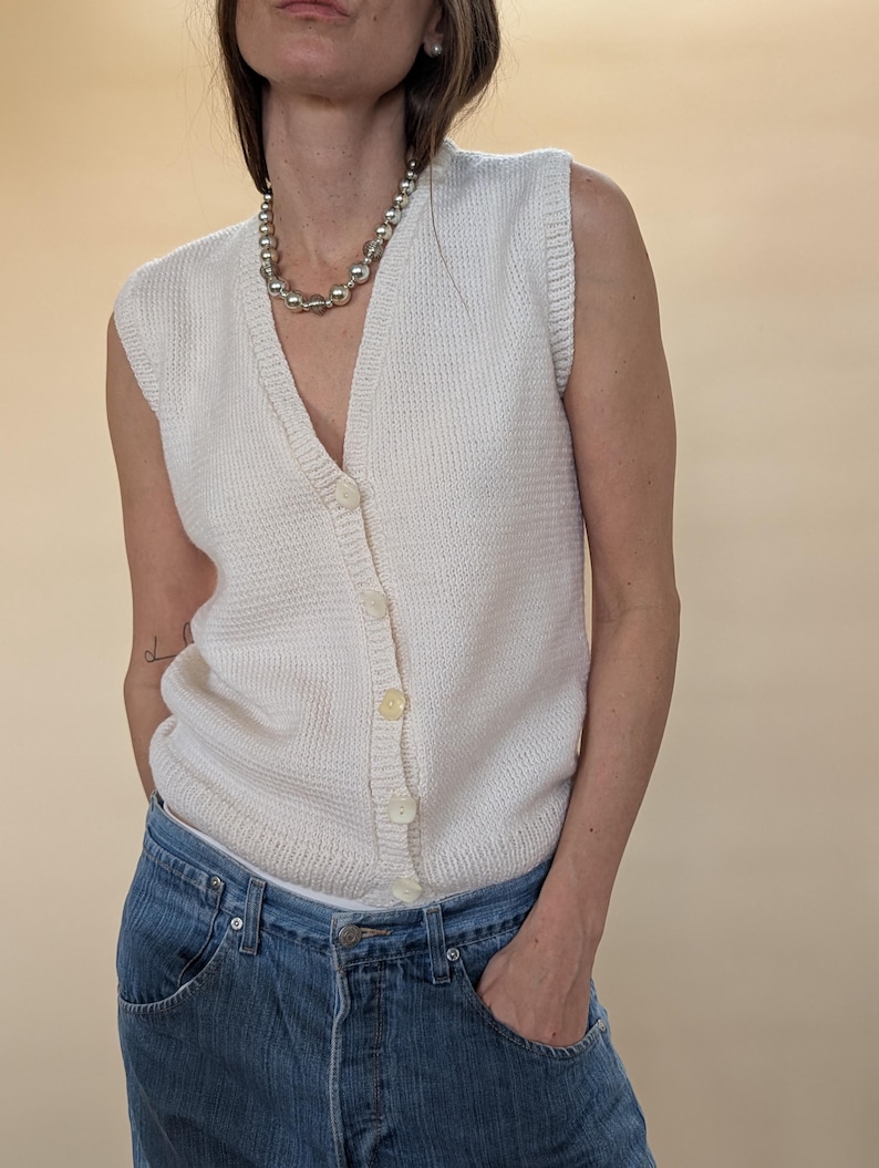 Peut inclure: Gilet sans manches cr&egrave;me avec un col en V et une fermeture boutonn&eacute;e. Le gilet est en tricot textur&eacute; et est associ&eacute; &agrave; un jean bleu. Un collier de perles argent&eacute;es est &eacute;galement visible.
