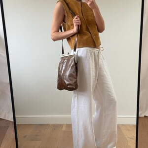 May include: A brown leather shoulder bag with a silver clasp, paired with a tan suede vest and white linen pants. The bag has a textured appearance and a long, thin strap. The vest has a button-up front.