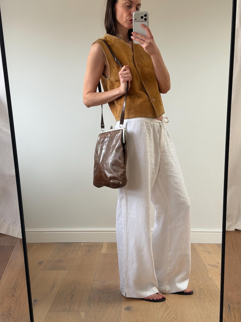 May include: A brown leather shoulder bag with a silver clasp, paired with a tan suede vest and white linen pants. The bag has a textured appearance and a long, thin strap. The vest has a button-up front.