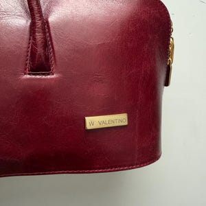 May include: A burgundy leather handbag with a top handle and a gold-toned zipper pull. The bag features a rectangular gold-toned plaque with the text "W VALENTINO". The leather has a textured appearance.