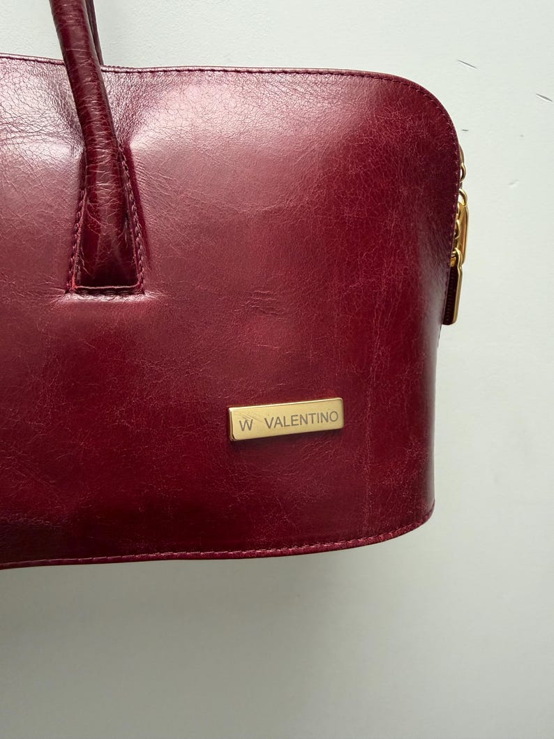 May include: A burgundy leather handbag with a top handle and a gold-toned zipper pull. The bag features a rectangular gold-toned plaque with the text "W VALENTINO". The leather has a textured appearance.
