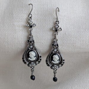 May include: A pair of silver-toned drop earrings. Each earring features a cameo portrait in a black oval frame, surrounded by small white beads and black accents. A black bead dangles from the bottom. The earrings have a decorative cross-shaped top.