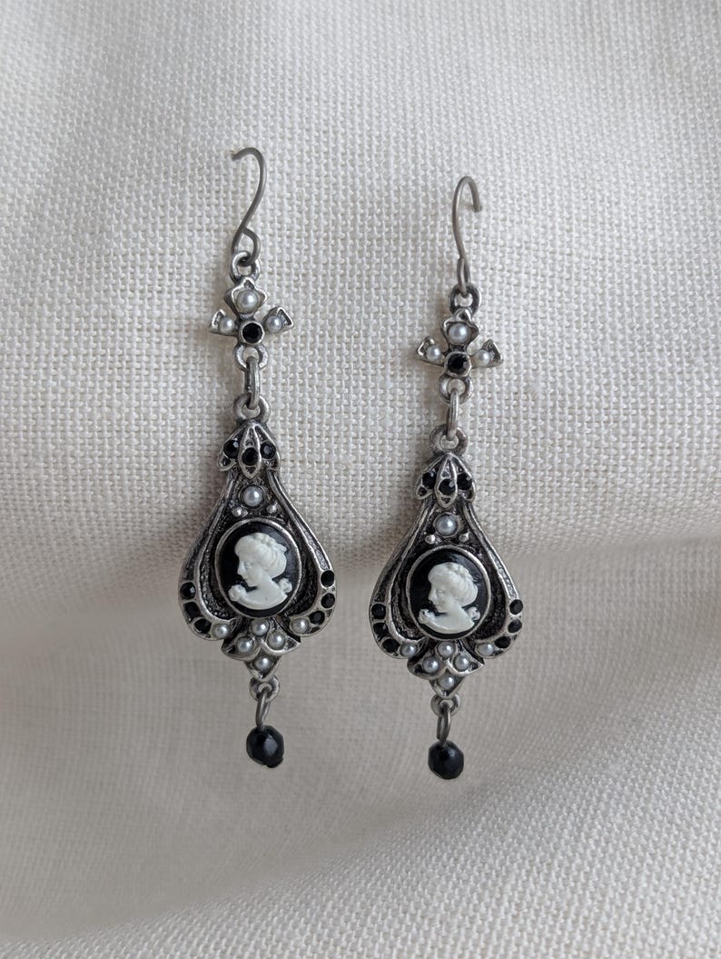 May include: A pair of silver-toned drop earrings. Each earring features a cameo portrait in a black oval frame, surrounded by small white beads and black accents. A black bead dangles from the bottom. The earrings have a decorative cross-shaped top.