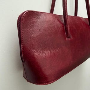 May include: A burgundy leather handbag with a rectangular shape and a zipper closure. The bag has two long, thin handles and a smooth, glossy finish. The bag is a deep red color.