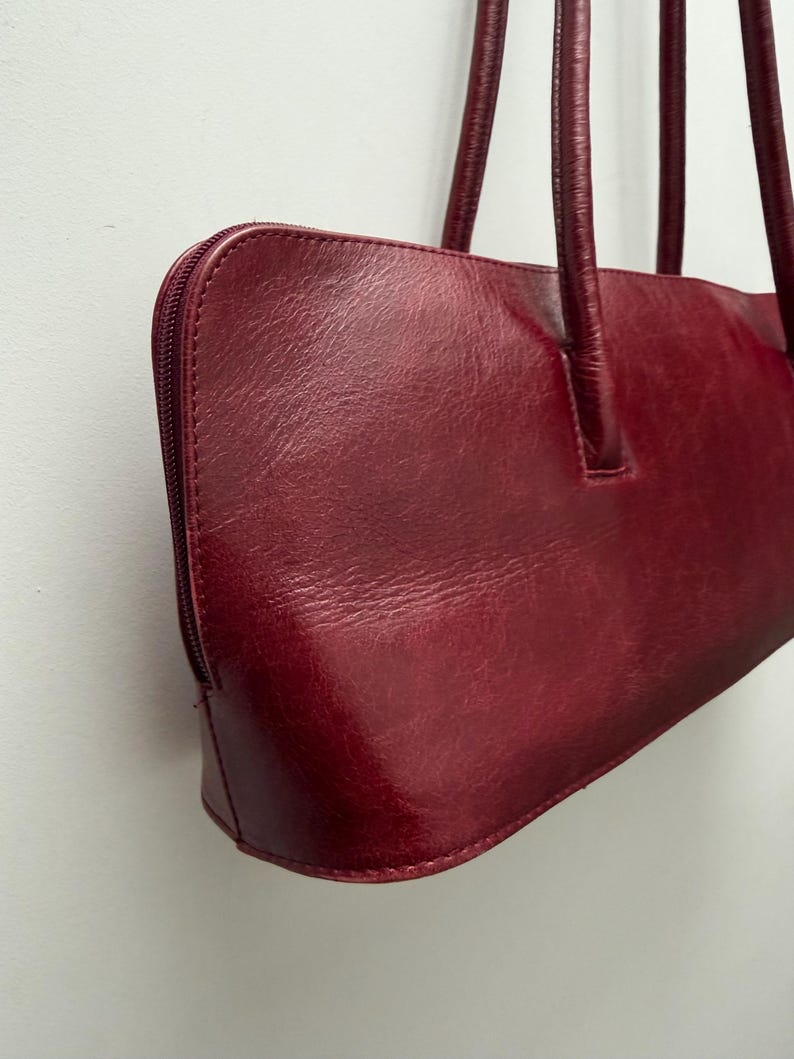 May include: A burgundy leather handbag with a rectangular shape and a zipper closure. The bag has two long, thin handles and a smooth, glossy finish. The bag is a deep red color.