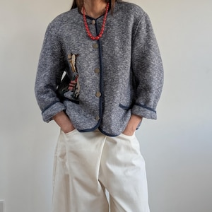 Könnte beinhalten: Eine blau-grau melierte Jacke mit dunkelblauen Paspeln und goldenen Knöpfen. Die Jacke hat ein gesticktes Design auf der Vorderseite und wird mit einer cremefarbenen Hose und einer roten Perlenkette kombiniert. Die Jacke hat zwei Taschen.