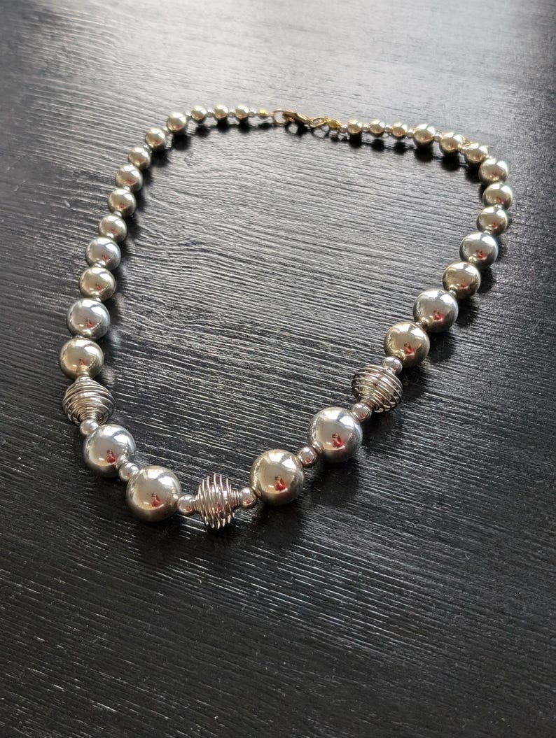 Peut inclure: Un collier argent&eacute; compos&eacute; de grosses perles rondes et d'&eacute;l&eacute;ments en fil de fer. Le collier est dot&eacute; d'un fermoir dor&eacute; et est pr&eacute;sent&eacute; sur une surface sombre. Ce bijou convient &agrave; diverses occasions. Longueur approximative : 40 cm.