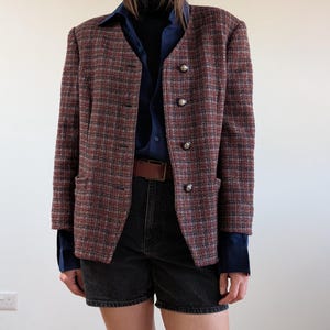 May include: A tweed blazer in shades of red, gray, and blue, featuring a classic plaid pattern. The blazer has a collarless design with three decorative silver buttons. The outfit includes a navy blue shirt, black denim shorts, and a brown belt.