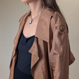 May include: A copper-colored trench coat with a large collar and button detail. The coat is worn over a black top and a necklace with a cameo pendant. Silver earrings are also visible.