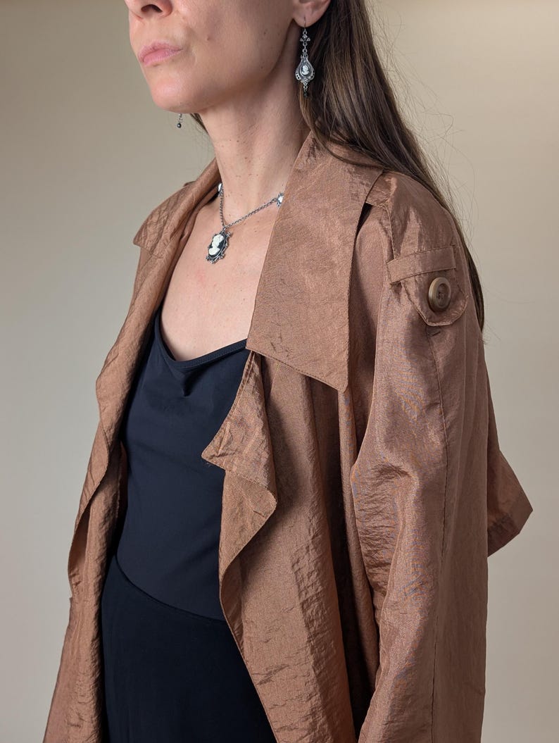 May include: A copper-colored trench coat with a large collar and button detail. The coat is worn over a black top and a necklace with a cameo pendant. Silver earrings are also visible.