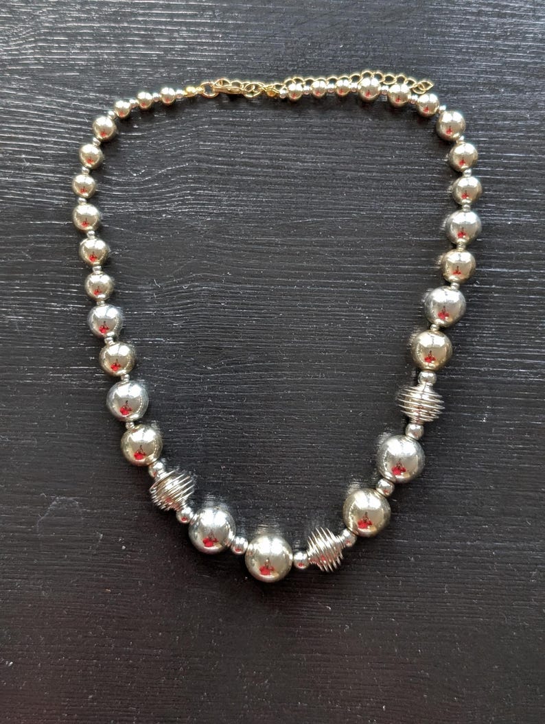 Peut inclure: Collier de perles argent&eacute;es avec fermoir &agrave; cha&icirc;ne dor&eacute;e. Le collier pr&eacute;sente un m&eacute;lange de grosses et petites perles rondes et brillantes argent&eacute;es, entrecoup&eacute;es de perles argent&eacute;es en forme de spirale.