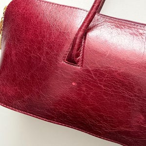 May include: A burgundy leather handbag with a rectangular shape and a single handle. The bag has a textured surface and a gold-colored zipper pull. The handle is a darker shade of burgundy.