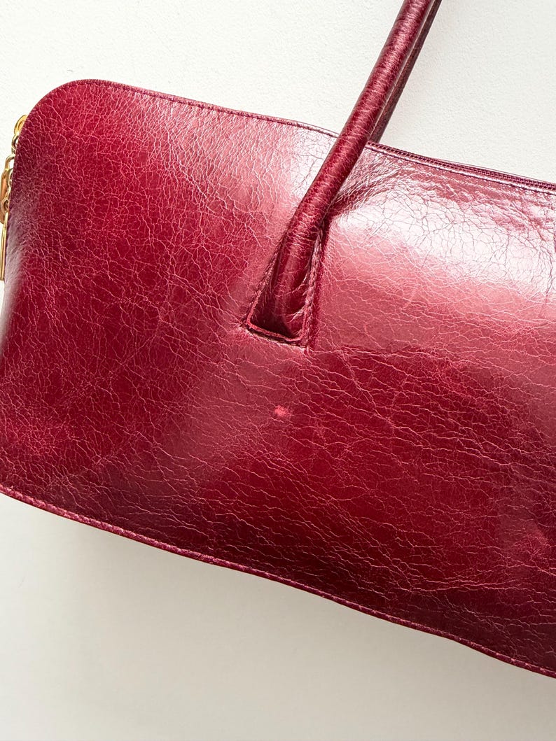 May include: A burgundy leather handbag with a rectangular shape and a single handle. The bag has a textured surface and a gold-colored zipper pull. The handle is a darker shade of burgundy.
