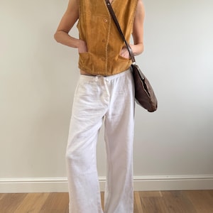May include: A tan suede vest with button closures and two front pockets is paired with white linen pants. A brown leather shoulder bag and black sandals complete the outfit. The clothing is casual and suitable for warm weather.