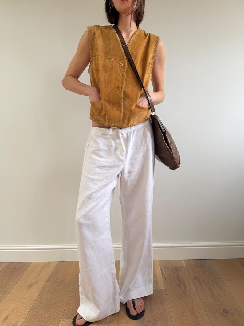 May include: A tan suede vest with button closures and two front pockets is paired with white linen pants. A brown leather shoulder bag and black sandals complete the outfit. The clothing is casual and suitable for warm weather.