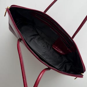 May include: Open burgundy leather tote bag with a black interior. The bag has two handles and a zippered closure. The words "Made in Italy" are visible inside the bag.