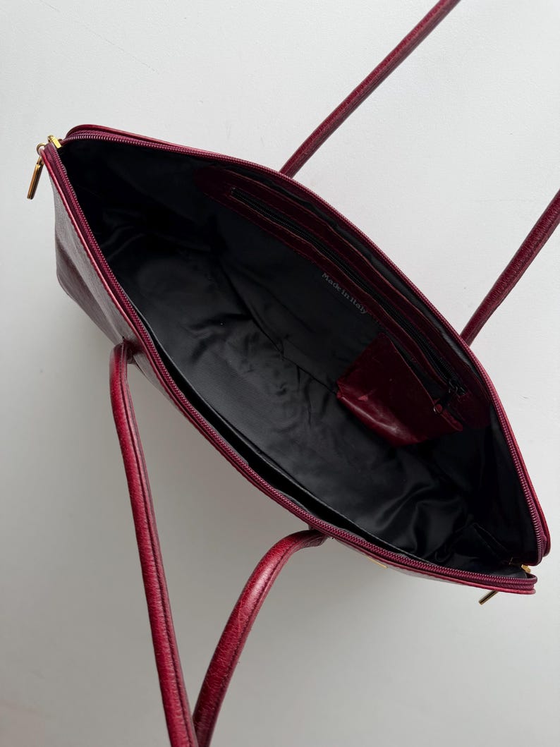 May include: Open burgundy leather tote bag with a black interior. The bag has two handles and a zippered closure. The words "Made in Italy" are visible inside the bag.