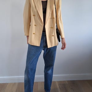 Op de afbeelding: Een lichtgele blazer met gouden knopen, gecombineerd met een blauwe jeans en zwarte hakken. De dubbelbreasted blazer wordt gecombineerd met een zwarte schoudertas. De outfit wordt gepresenteerd tegen een witte muur en een houten vloer.