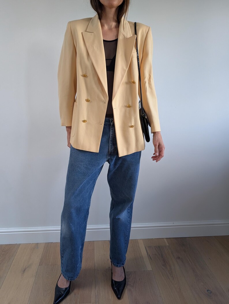 Op de afbeelding: Een lichtgele blazer met gouden knopen, gecombineerd met een blauwe jeans en zwarte hakken. De dubbelbreasted blazer wordt gecombineerd met een zwarte schoudertas. De outfit wordt gepresenteerd tegen een witte muur en een houten vloer.