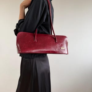May include: A rectangular, red handbag with long handles. The bag has a smooth, glossy finish and a small, rectangular gold-colored accent near the top. The bag is carried over the shoulder. The overall style is classic and elegant.