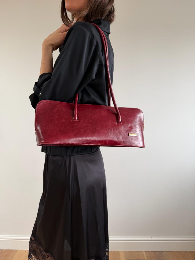 May include: A rectangular, red handbag with long handles. The bag has a smooth, glossy finish and a small, rectangular gold-colored accent near the top. The bag is carried over the shoulder. The overall style is classic and elegant.