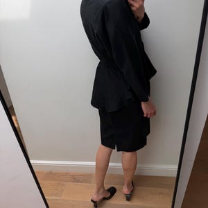 Puede incluir: Un conjunto de dos piezas negro que consta de una chaqueta de manga larga con cintur&oacute;n en la cintura y una falda a juego hasta la rodilla. La chaqueta presenta un cuello y un dobladillo estilo peplum, mientras que la falda tiene un corte recto con una peque&ntilde;a abertura lateral.