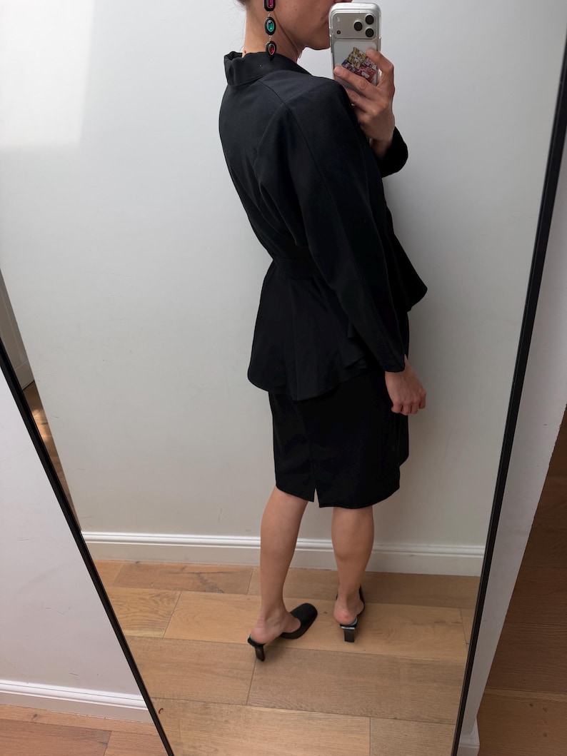 Puede incluir: Un conjunto de dos piezas negro que consta de una chaqueta de manga larga con cintur&oacute;n en la cintura y una falda a juego hasta la rodilla. La chaqueta presenta un cuello y un dobladillo estilo peplum, mientras que la falda tiene un corte recto con una peque&ntilde;a abertura lateral.