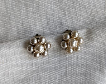 May include: A pair of pearl cluster earrings. Each earring features a flower-like design composed of multiple faux pearls. The earrings are set against a white fabric background, creating a classic and elegant aesthetic.