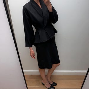 Puede incluir: Un conjunto negro de dos piezas compuesto por una chaqueta de manga larga con peplum y cuello chal, y una falda a juego a la altura de la rodilla. La chaqueta presenta una cintura entallada y un bajo acampanado, junto a una falda recta y mules negros.
